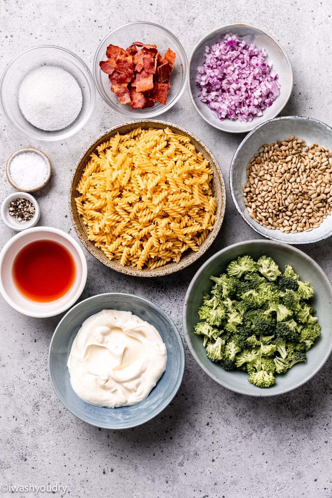 ingredients for broccoli pasta salad in small bowls arranged on white surface.