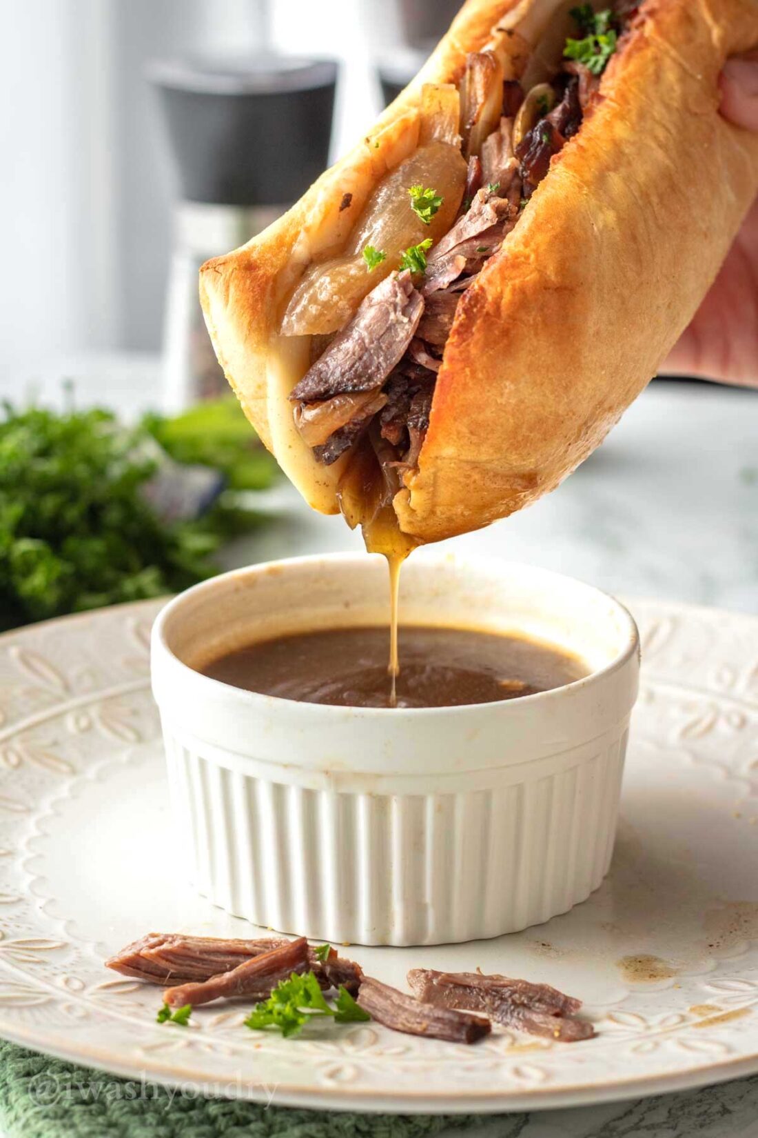 Cooked french dip sandwich dipping into white dish of au jus sauce. 