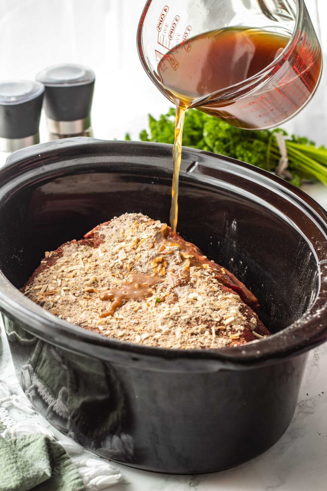 Pouring beef broth into black crock pot with raw chuck roast. 