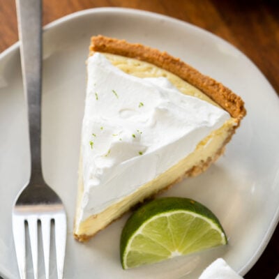 Slice of key lime pie on white plate with fork and lime slice.