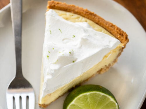 Slice of key lime pie on white plate with fork and lime slice.