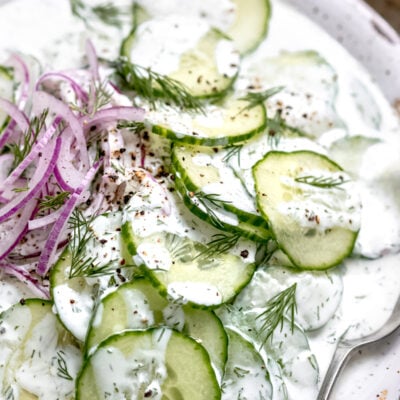 Cold sliced cucumbers and purple onions in a white creamy dressing with spoon in a white bowl.