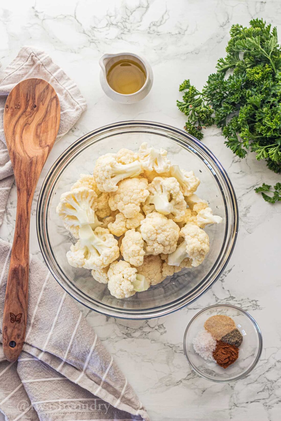 Chopped cauliflower florets, olive oil, and spices in glass bowls with wooden spoon. 