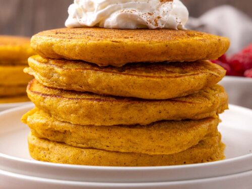 stack of pumpkin pancakes on white plate with whipped cream on top.