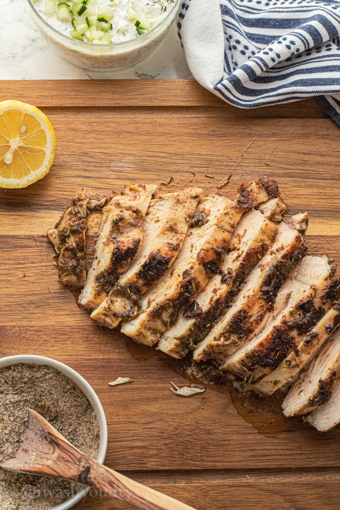 Sliced cooked chicken breast on wood board with white bowl of spice mix and lemon slice. 