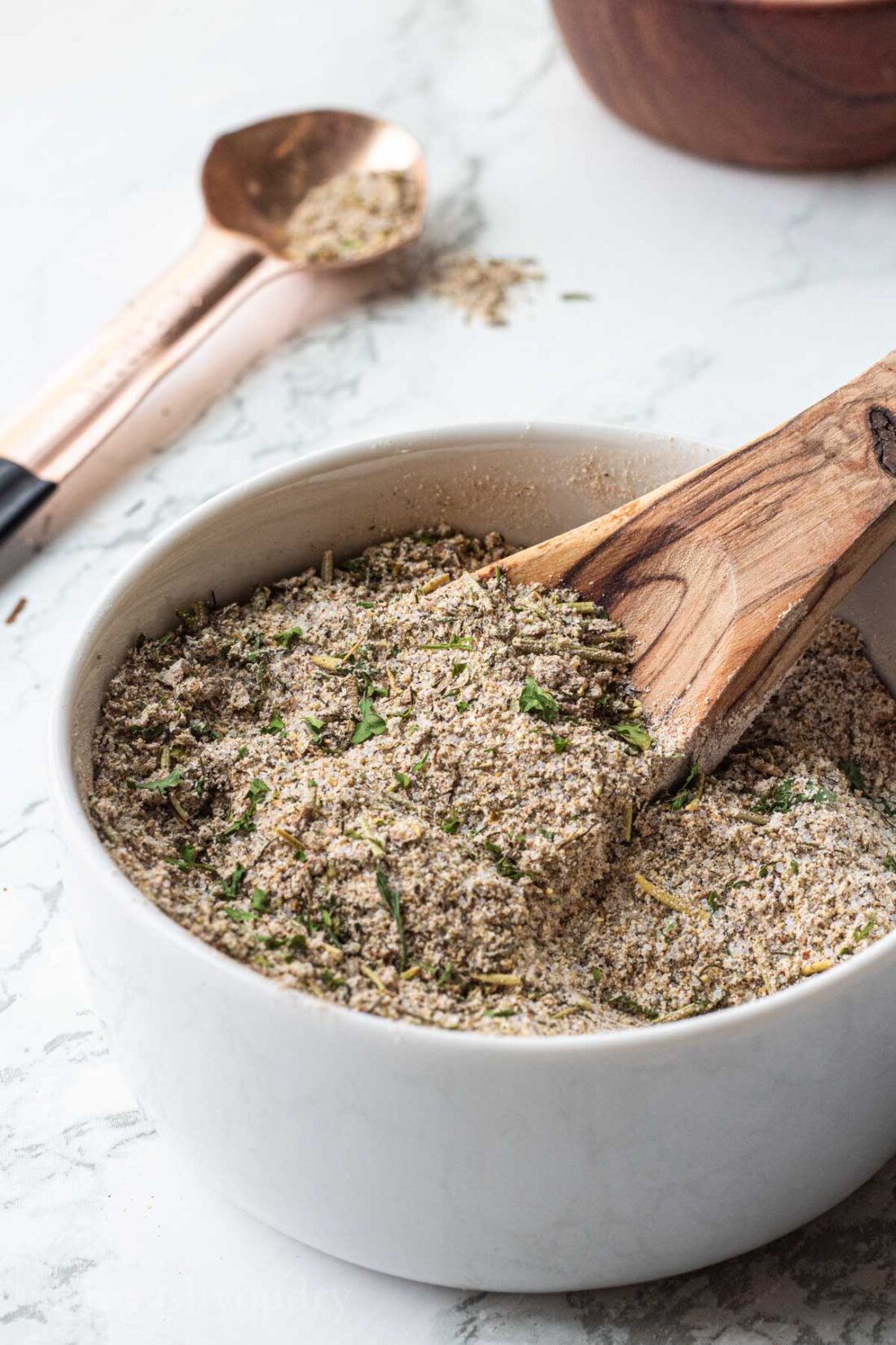 Greek spice blend with wooden spoon in white bowl.