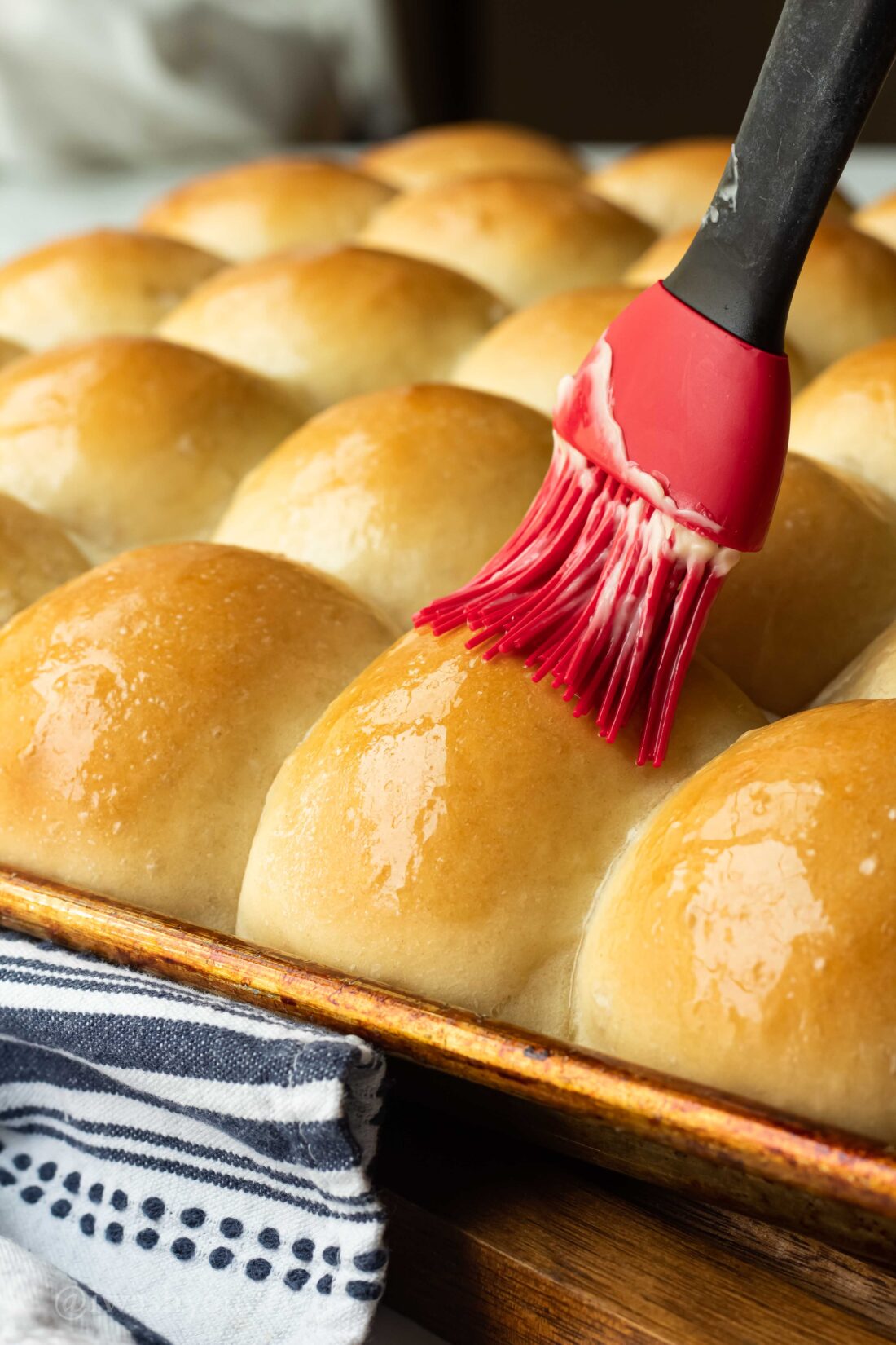 Red brush with butter over warm homemade rolls on metal pan. 