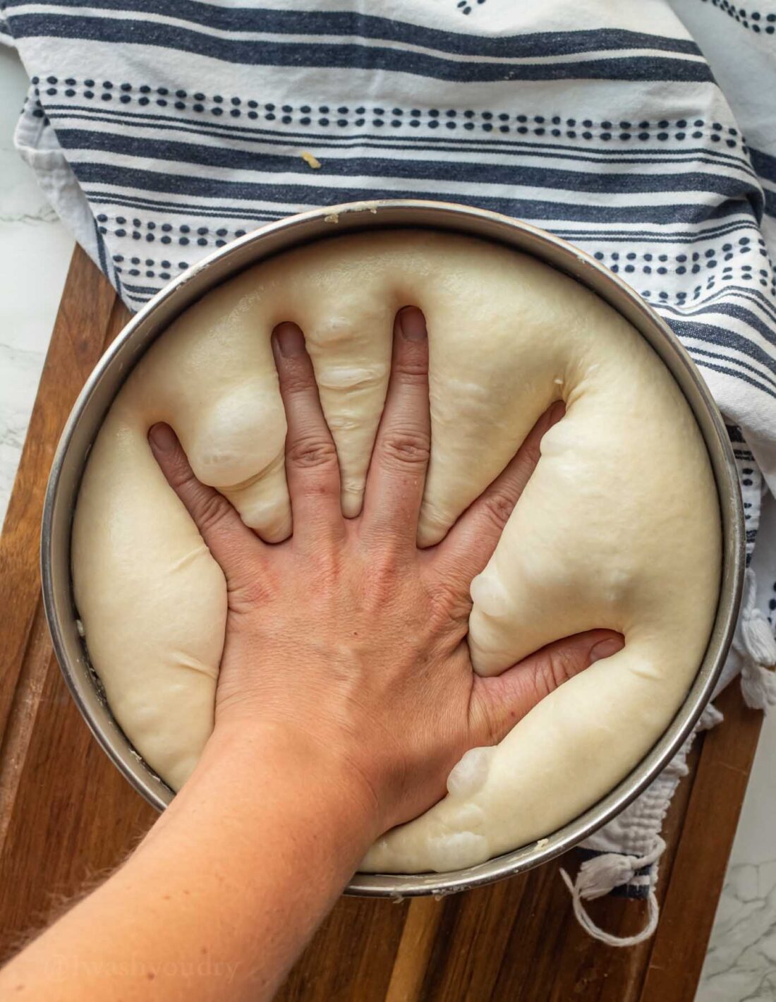Hand punching risen raw roll dough in metal mixing bowl. 