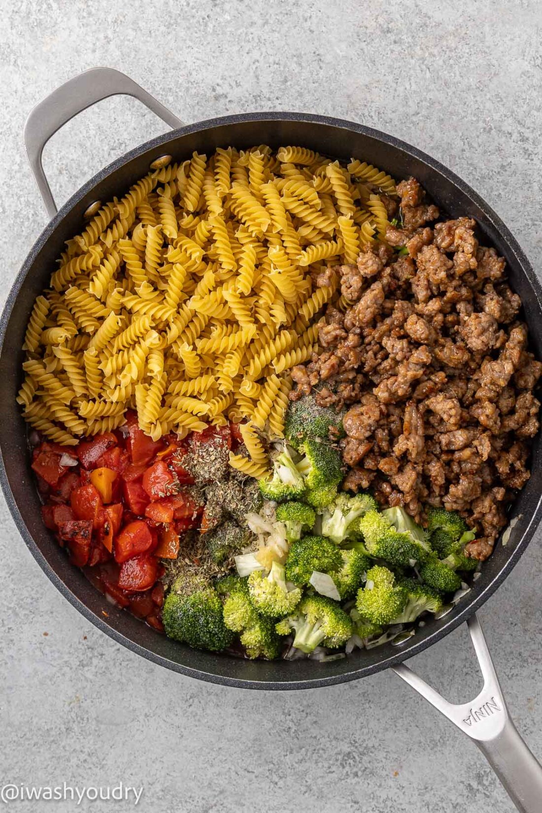 Noodles, tomatoes, broccoli, sausage, and spices in metal pan. 