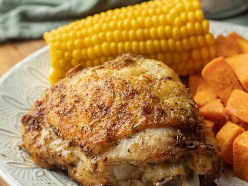 Cooked chicken thigh on white plate with corn and sweet potato.