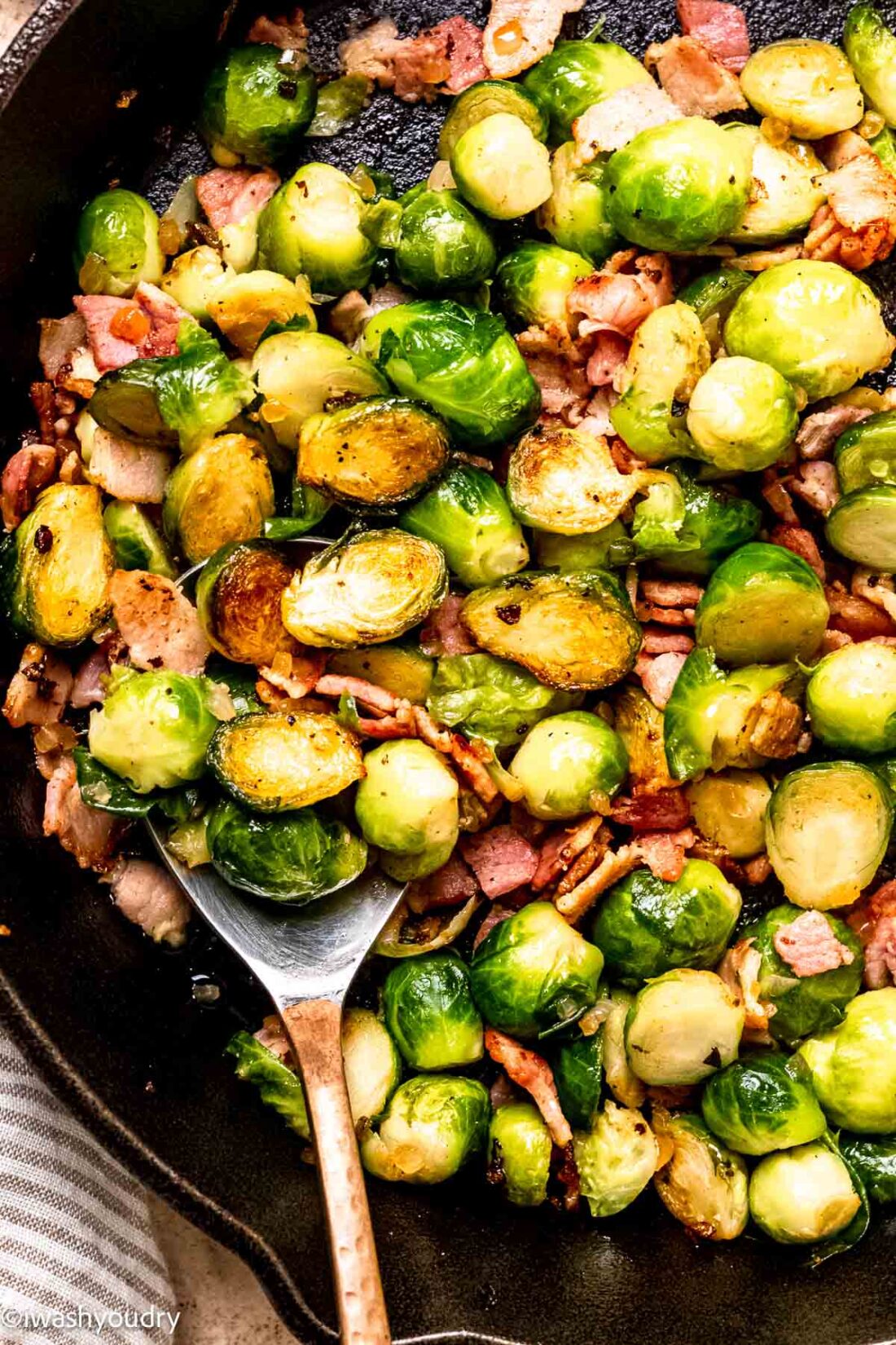 Cooked Brussels sprouts with bacon in black frying pan with spoon. 