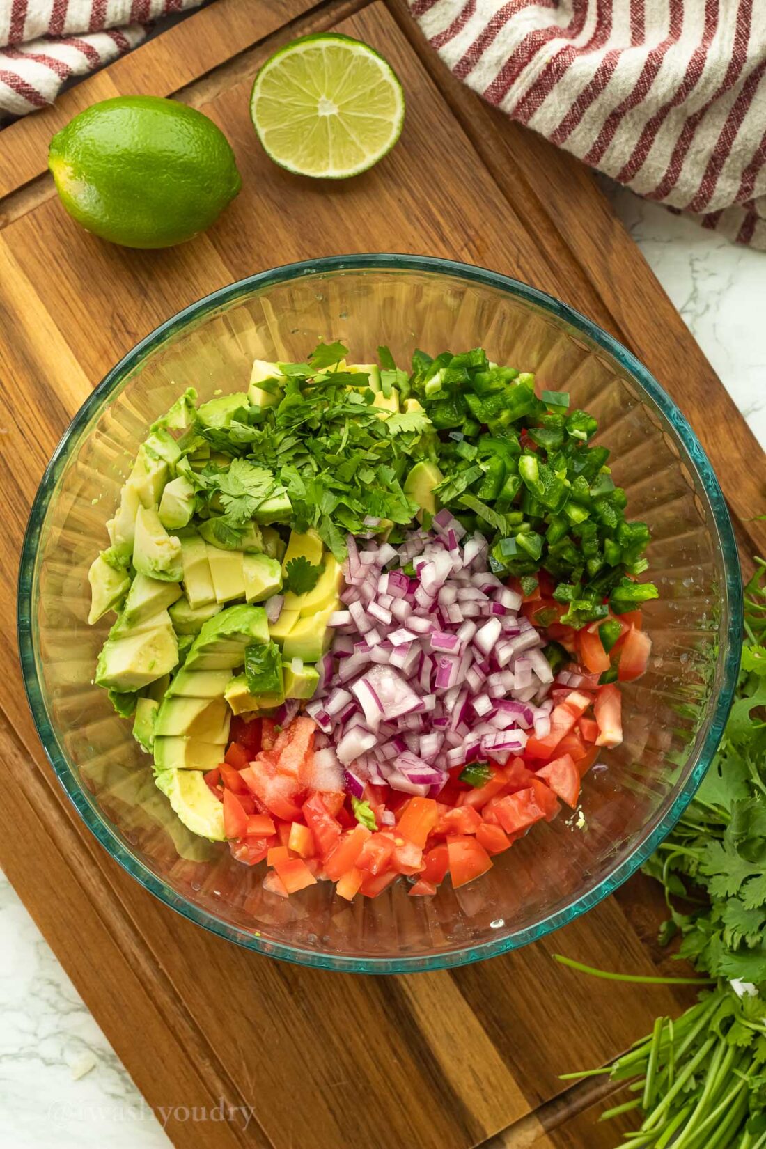 Avocado, jalapeno, purple onion, tomato, cilantro, and lime juice in glass bowl on wooden cutting board. 