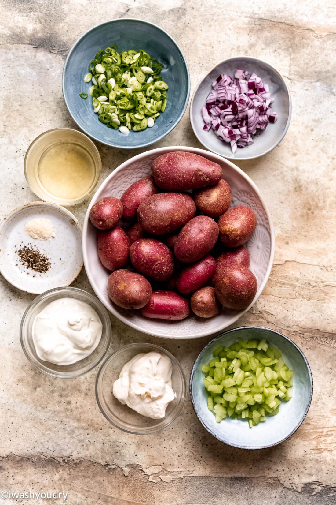 Ingredients for red potato salad in ceramic bowls on marble countertop. 
