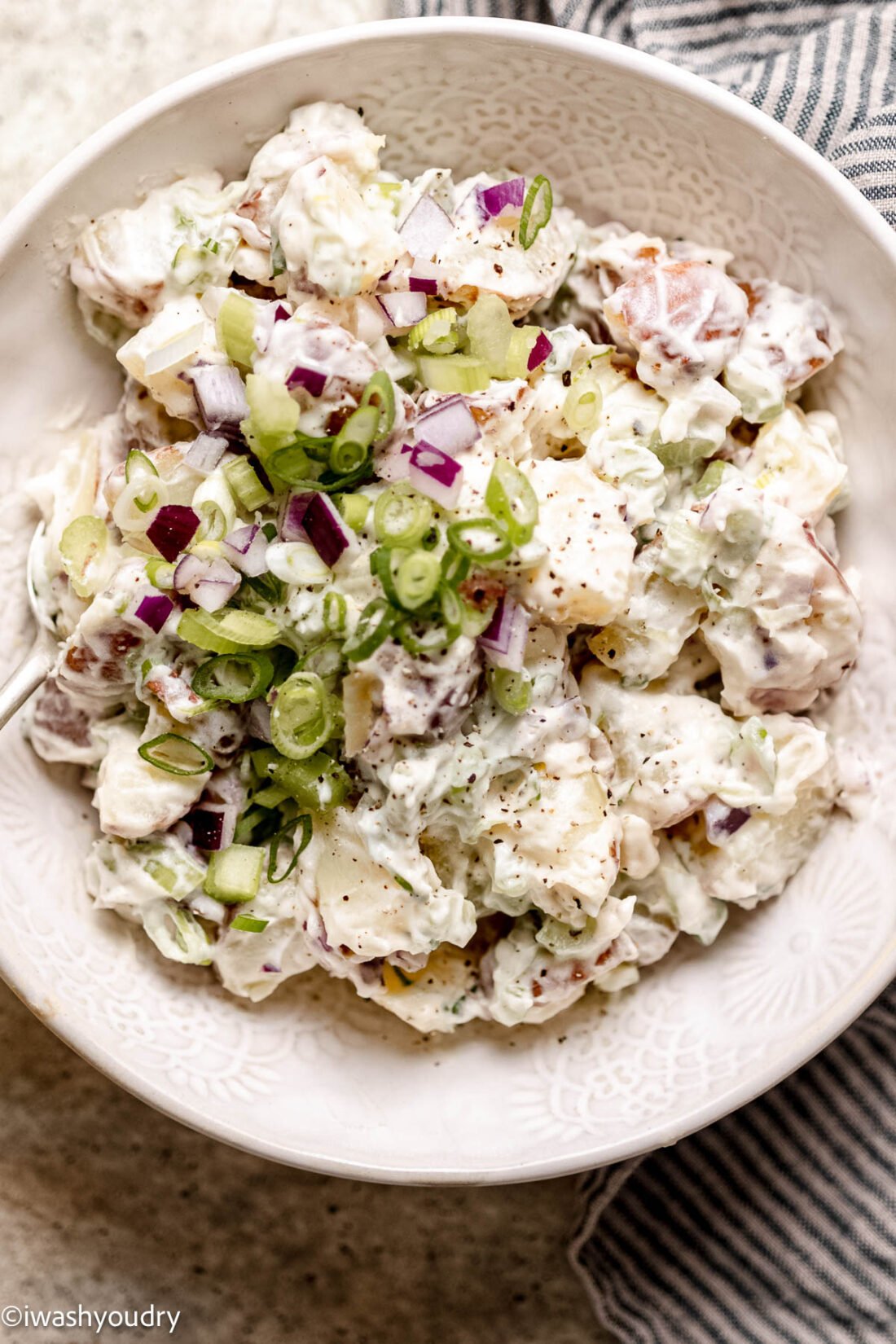 Creamy red potato salad in a white bowl with green onions and cracked pepper on top.