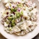 Creamy red potato salad in a white bowl with green onions and cracked pepper on top.