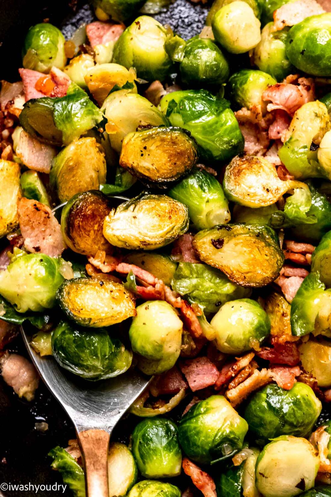 Brussels sprouts with bacon in black pan with spoon.