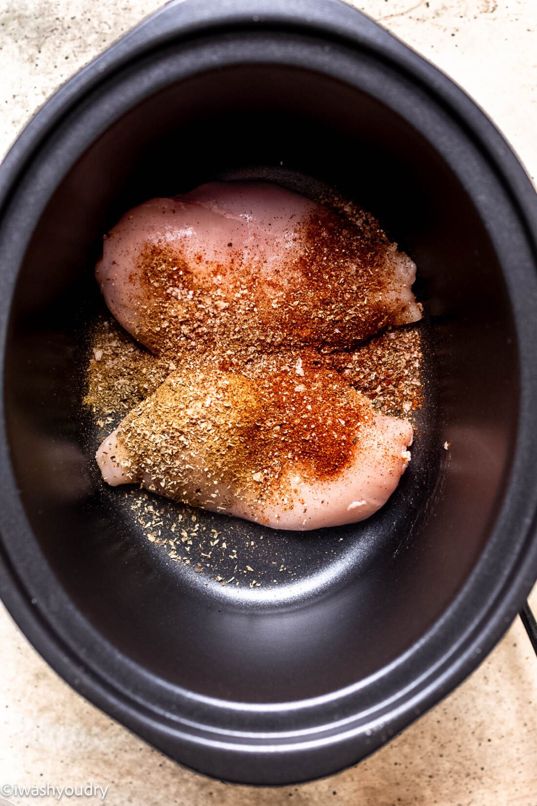 Raw chicken breasts in bowl of slow cooker with seasoning on top. 
