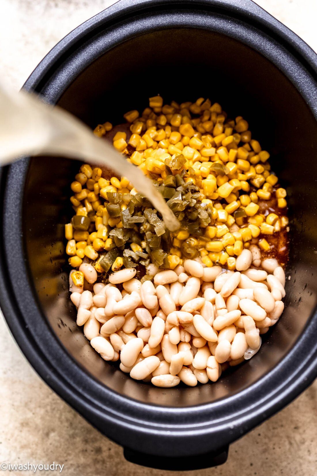 Pouring chicken broth on chicken corn, chiles, beans and seasoning in slow cooker bowl. 