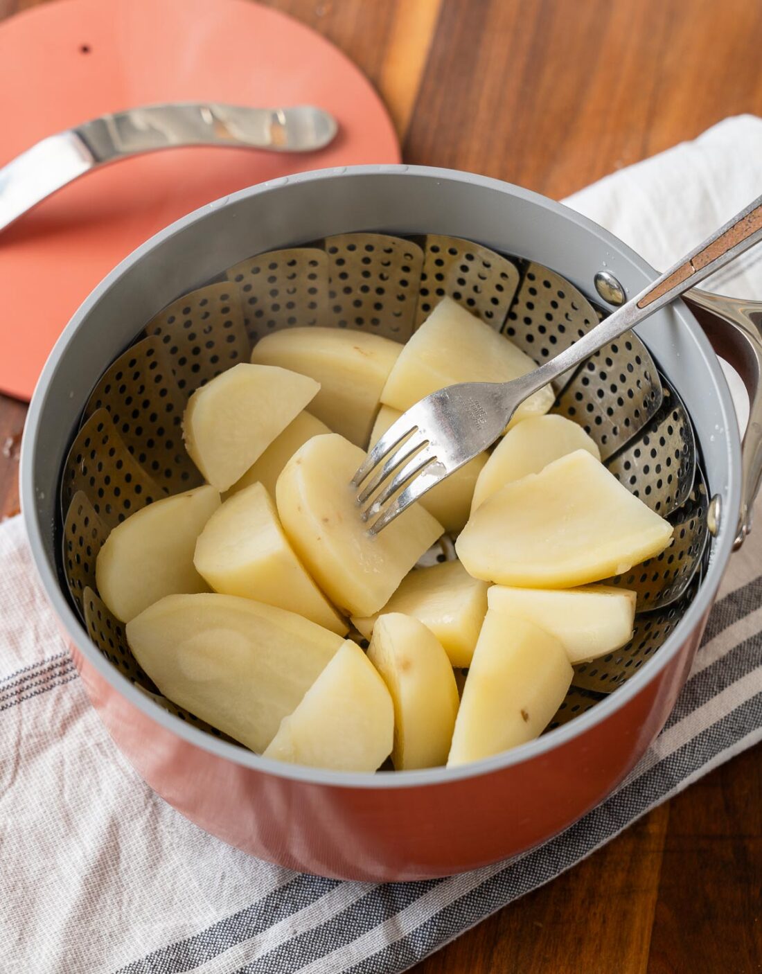 Fork piercing steamed quartered potatoes in steamer basket in red pot. 