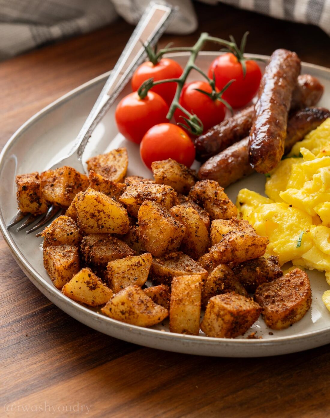 Cooked breaskfast skillet potatoes on white plate with tomatoes, sausages,a nd scrambled eggs and fork. 