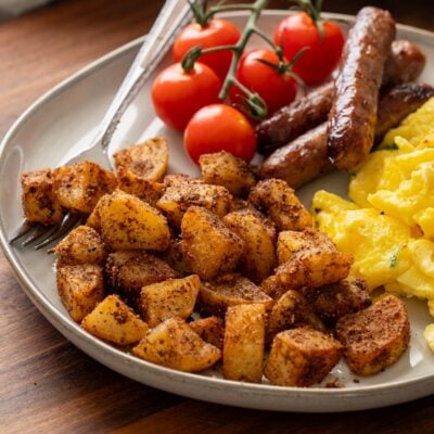 Cooked skillet breakfast potatoes on white plate with tomatoes. sausages, scrambled eggs, and a fork.