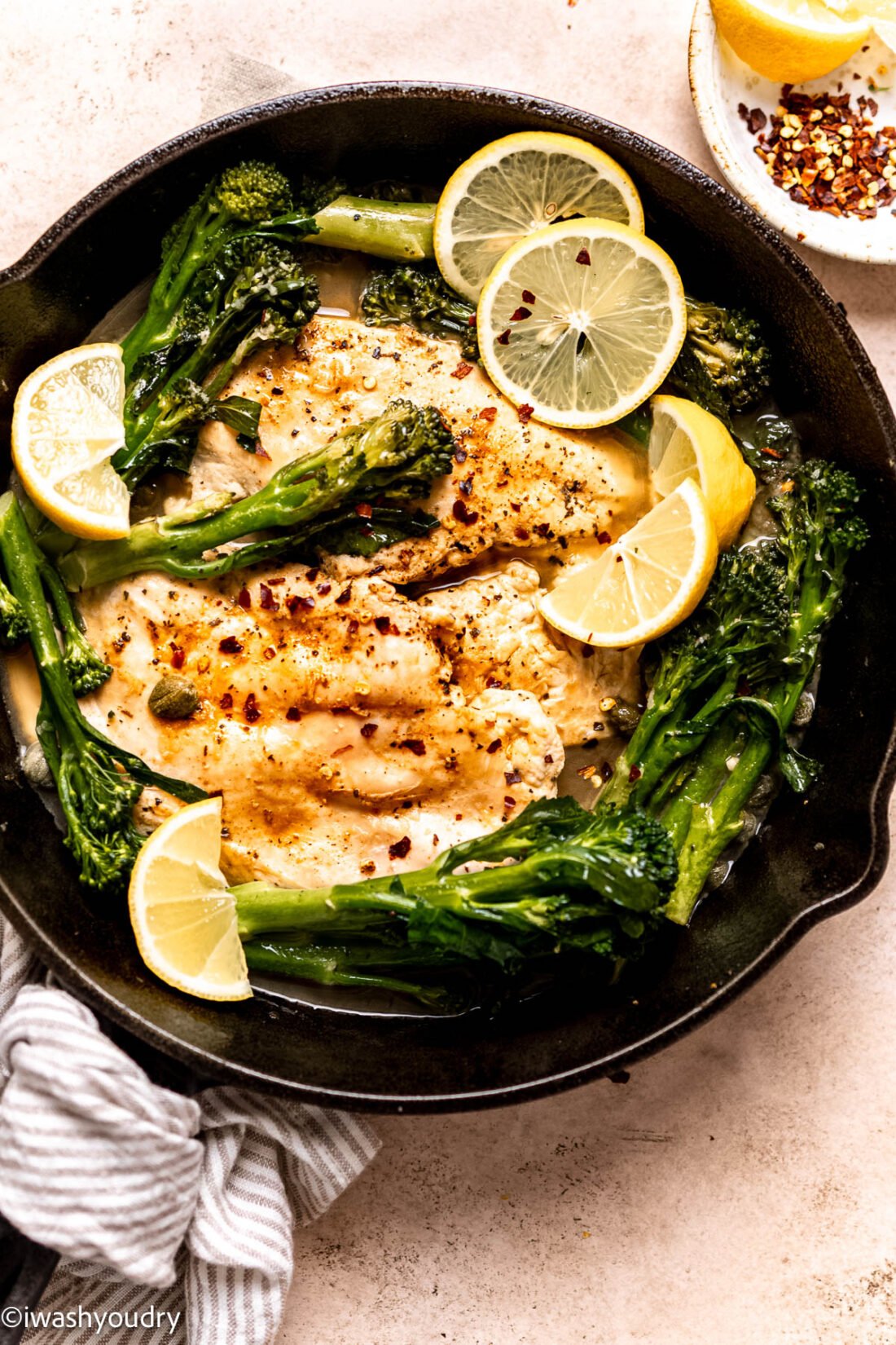 Cooked chicken and broccolini in frying pan with lemon wedges and capers. 