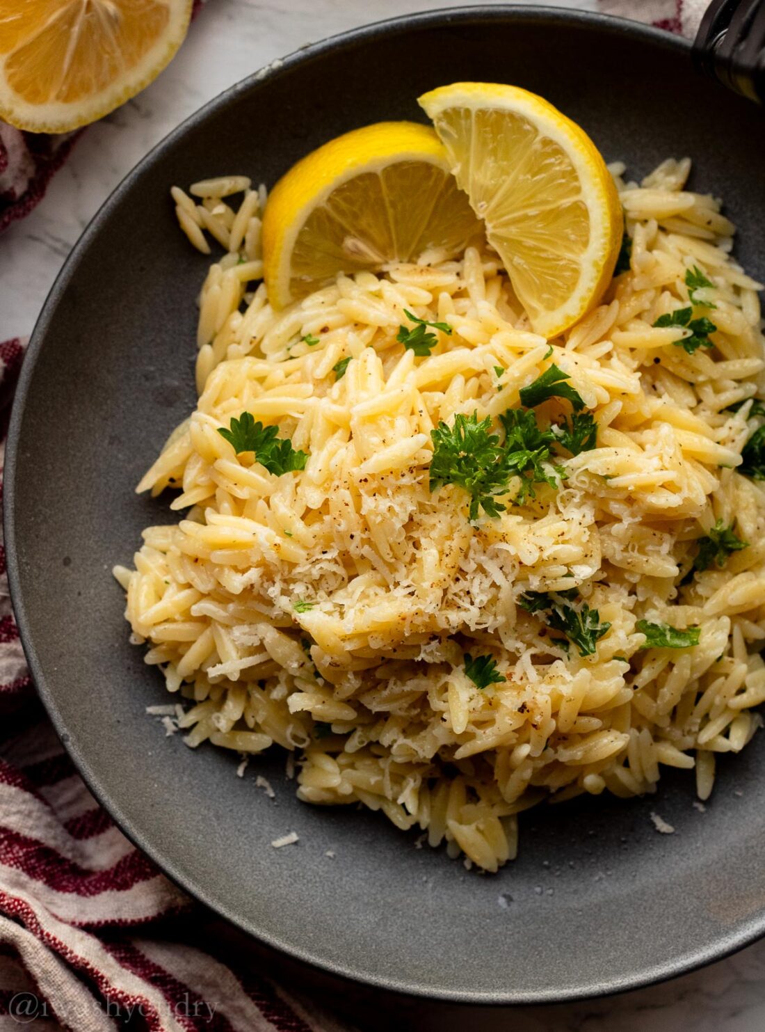 Cooked orzo pasta in black bowl with parsley and lemon wedge with dish towel and lemon half on the side. 