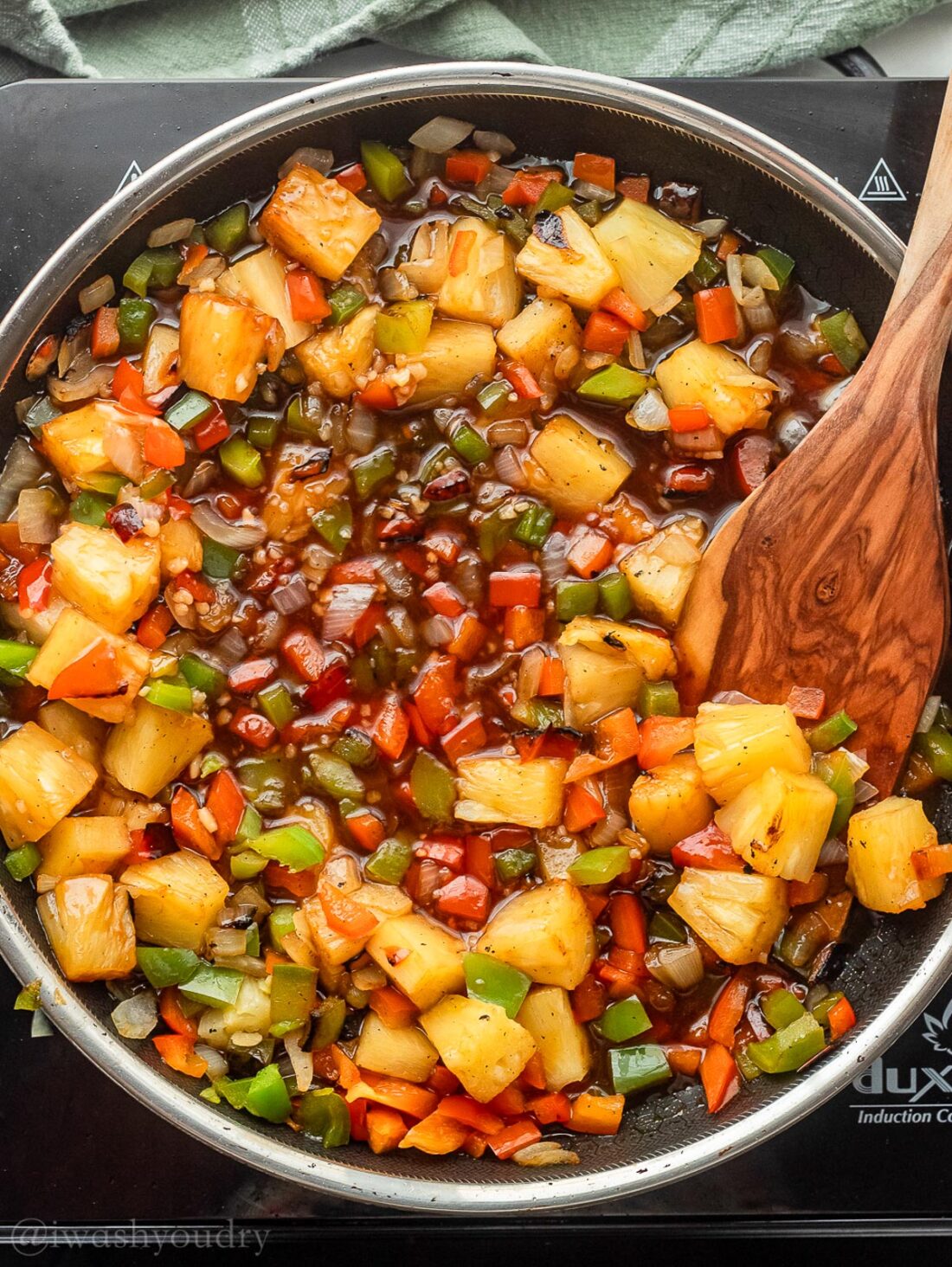 Pineapple and peppers in frying pan of sweet and sour sauce with wooden spatula. 
