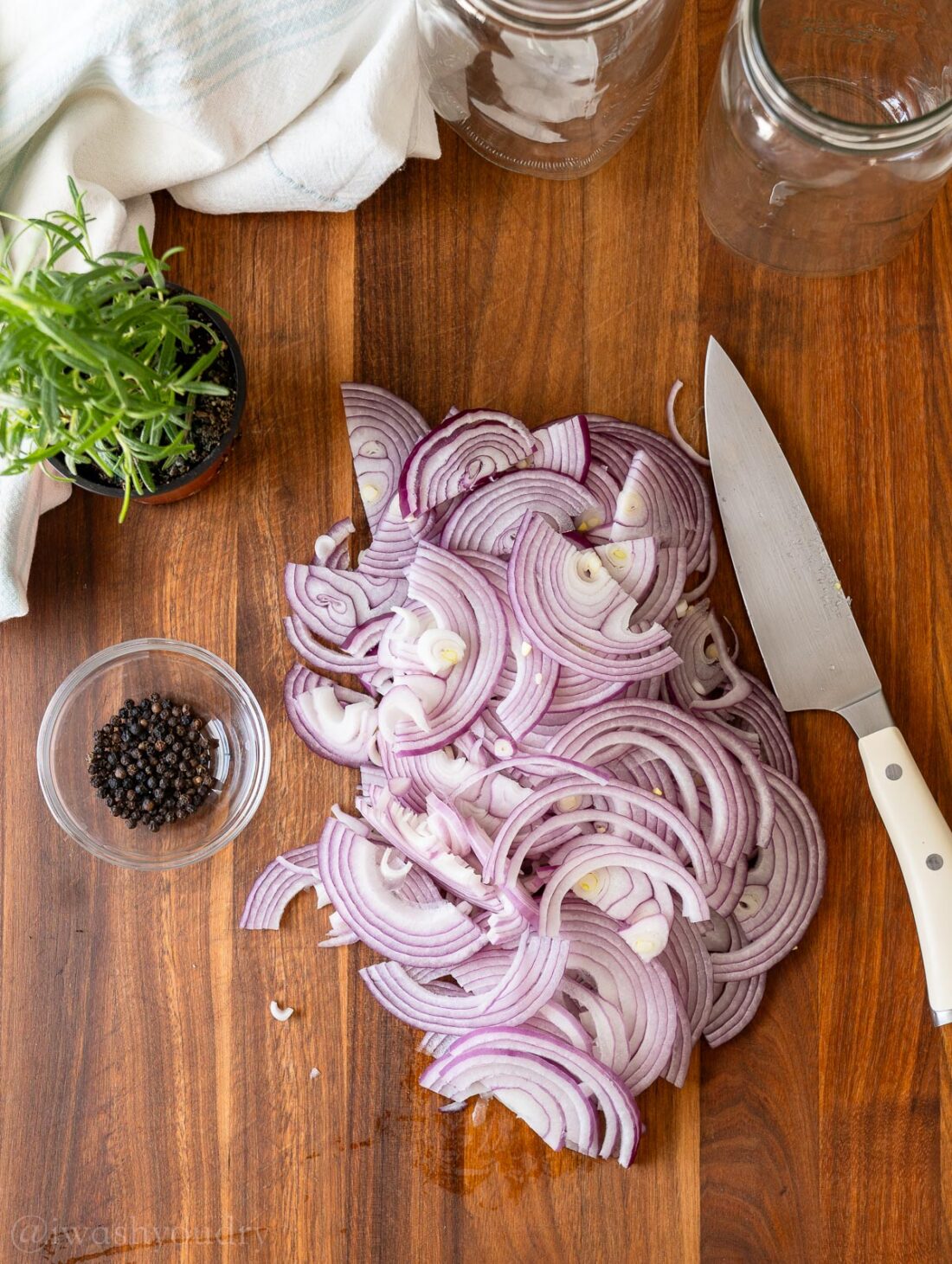 Sliced red onions on wood cutting board with rosemary and bowl of peppercorns. 
