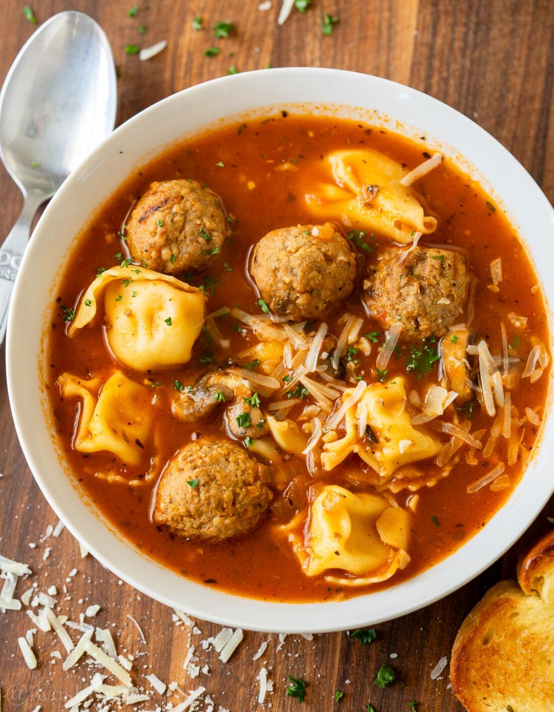 White bowl full of cooked meatball tortellini soup with spoon. 