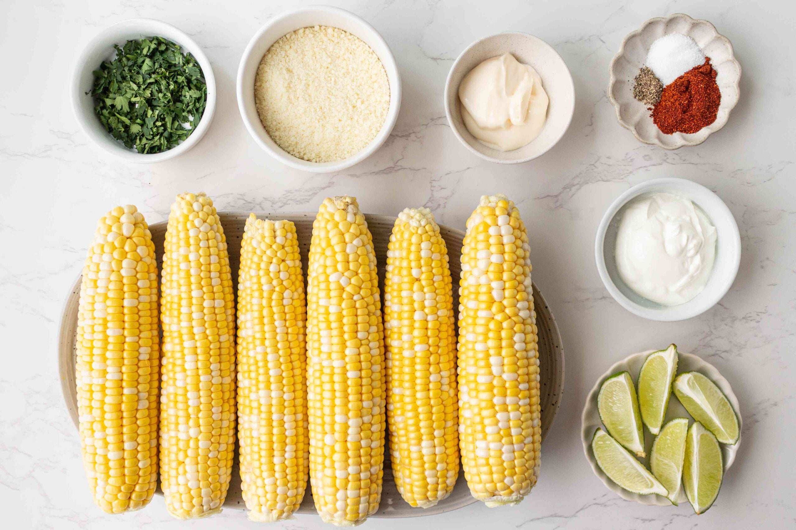 ingredients for elote street corn on white surface with seasonings and lime.