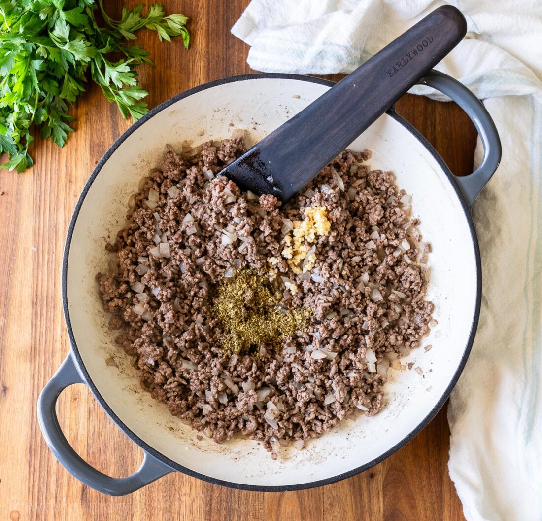 browned beef in pan with garlic and seasoning. wooden spoon sticking out of pan.