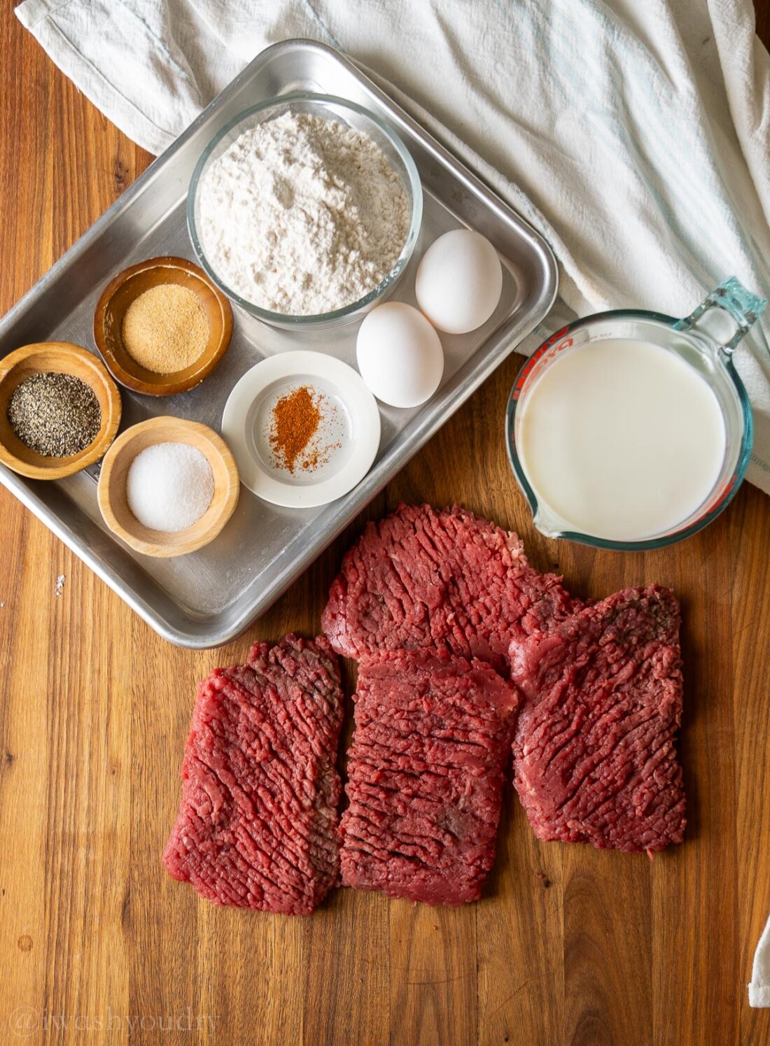 cube steaks on a wooden surface with ingredients like flour, eggs, milk and seasonings beside it.