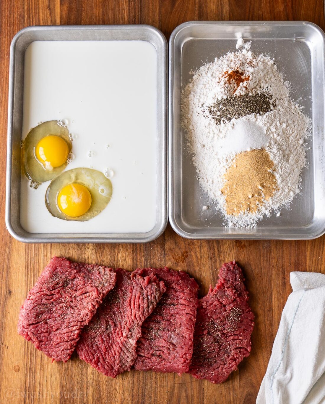 small sheet pans with breading in one pan, eggs and milk in another, cube steaks on wooden surface.