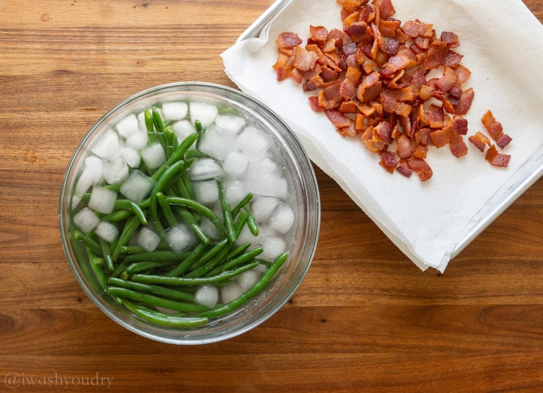 green beans in a bowl of ice water with crispy bacon in a pan next to it.
