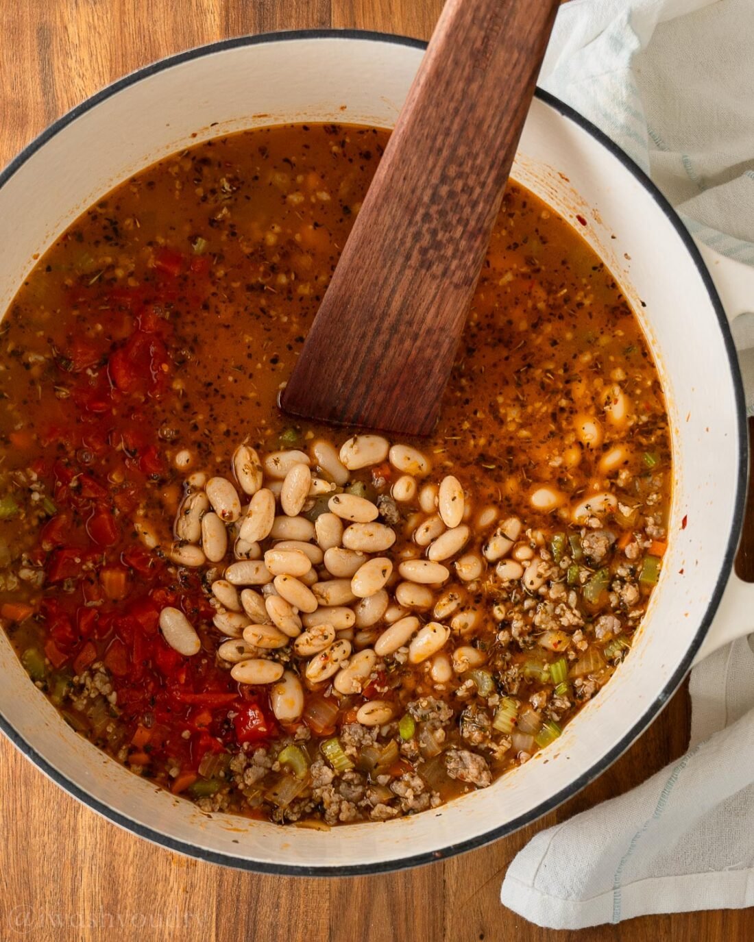 pot of soup with white beans and tomatoes with wooden spoon inside.