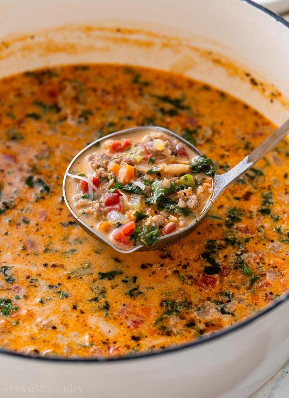 sausage and kale soup in a ladle inside of a white pot.