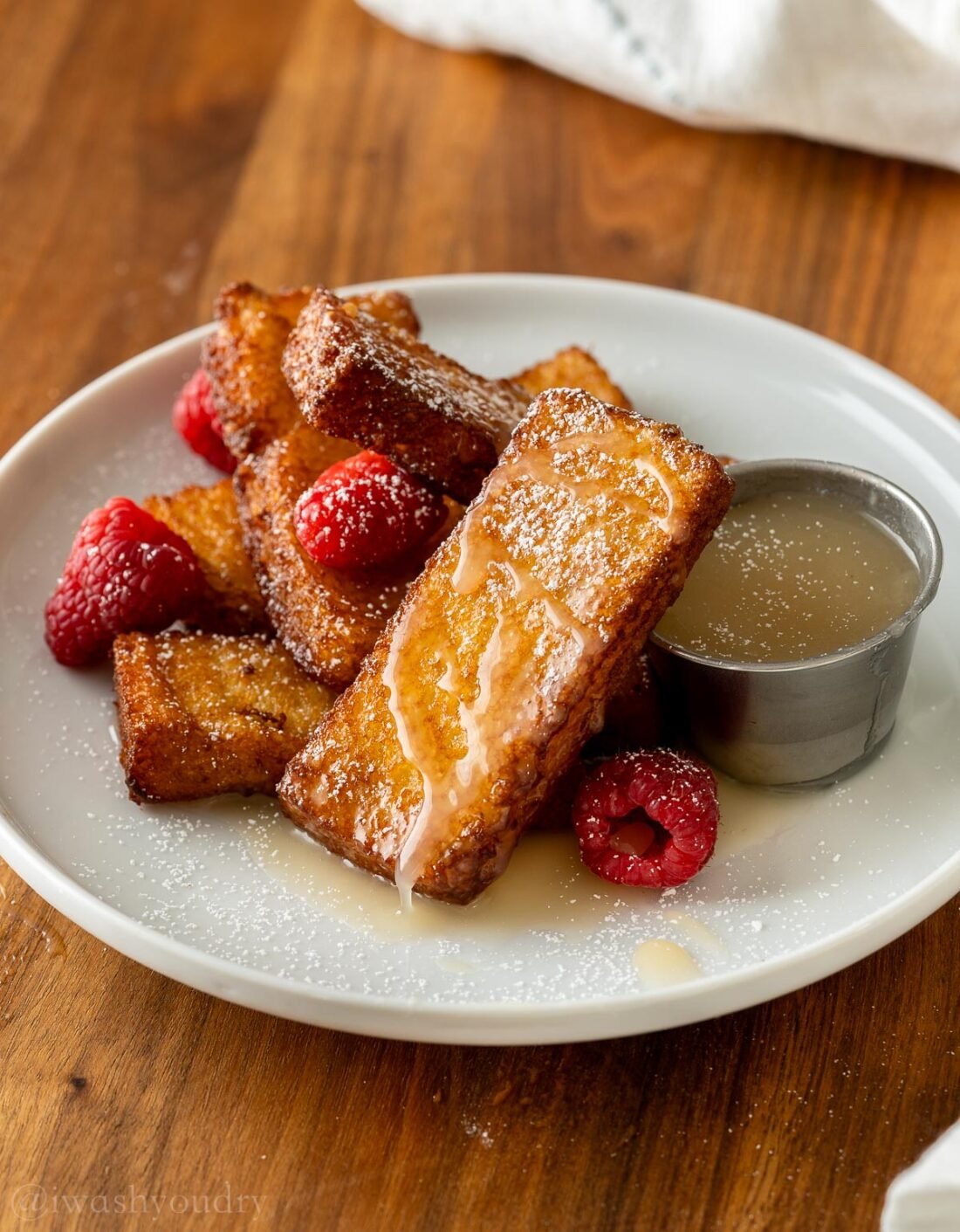 French Toast sticks stacked on a white plate with buttermilk syrup next to it