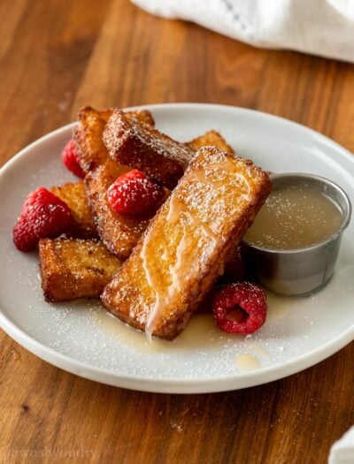 French Toast sticks stacked on a white plate with buttermilk syrup next to it