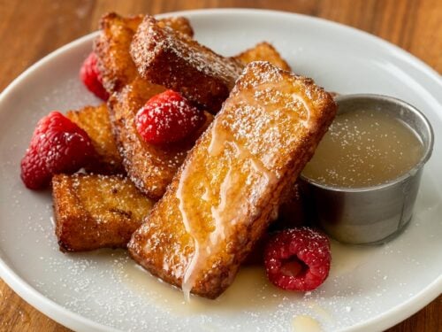 French Toast sticks stacked on a white plate with buttermilk syrup next to it