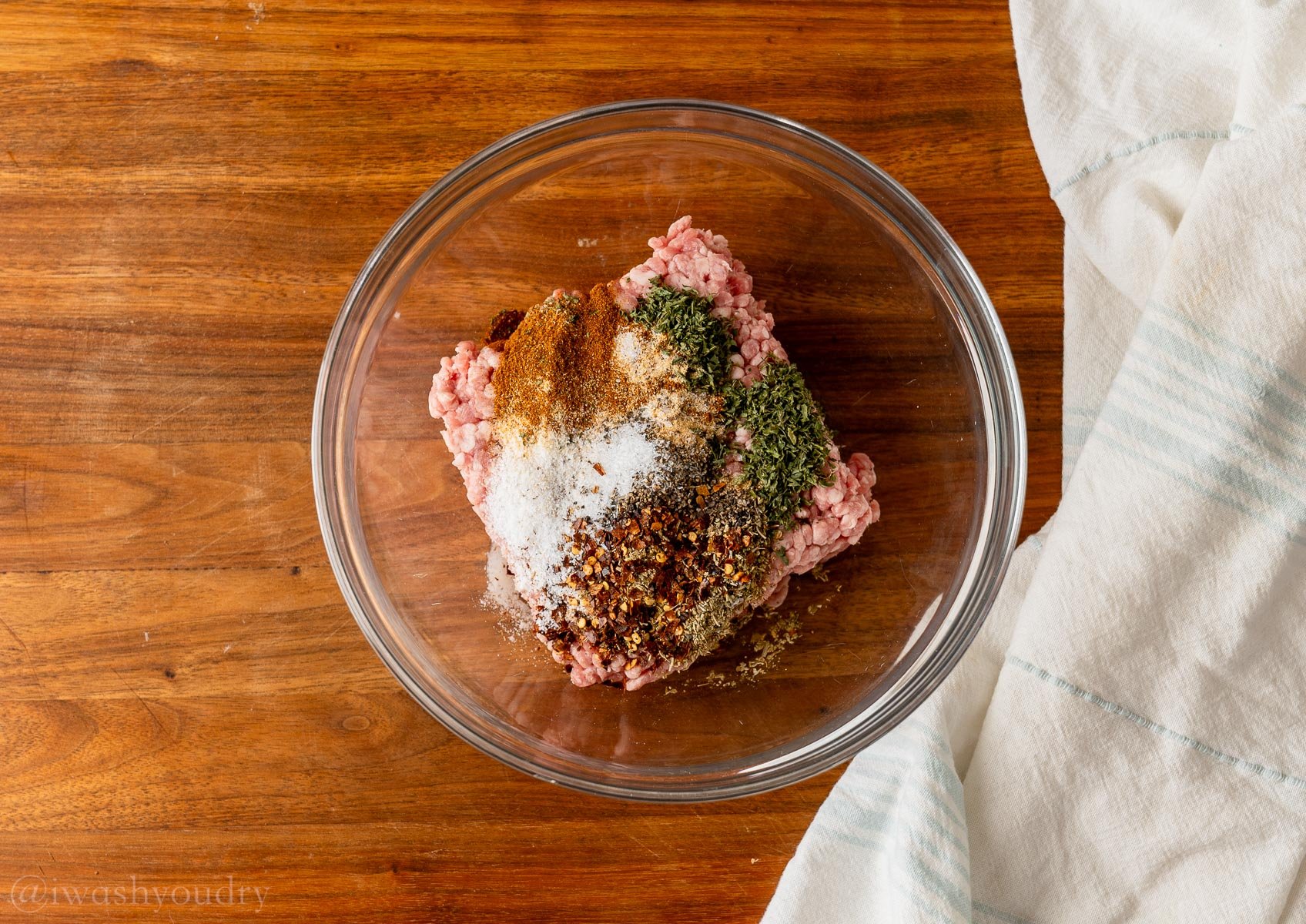 seasonings on top of ground pork in clear glass bowl on wooden surface.