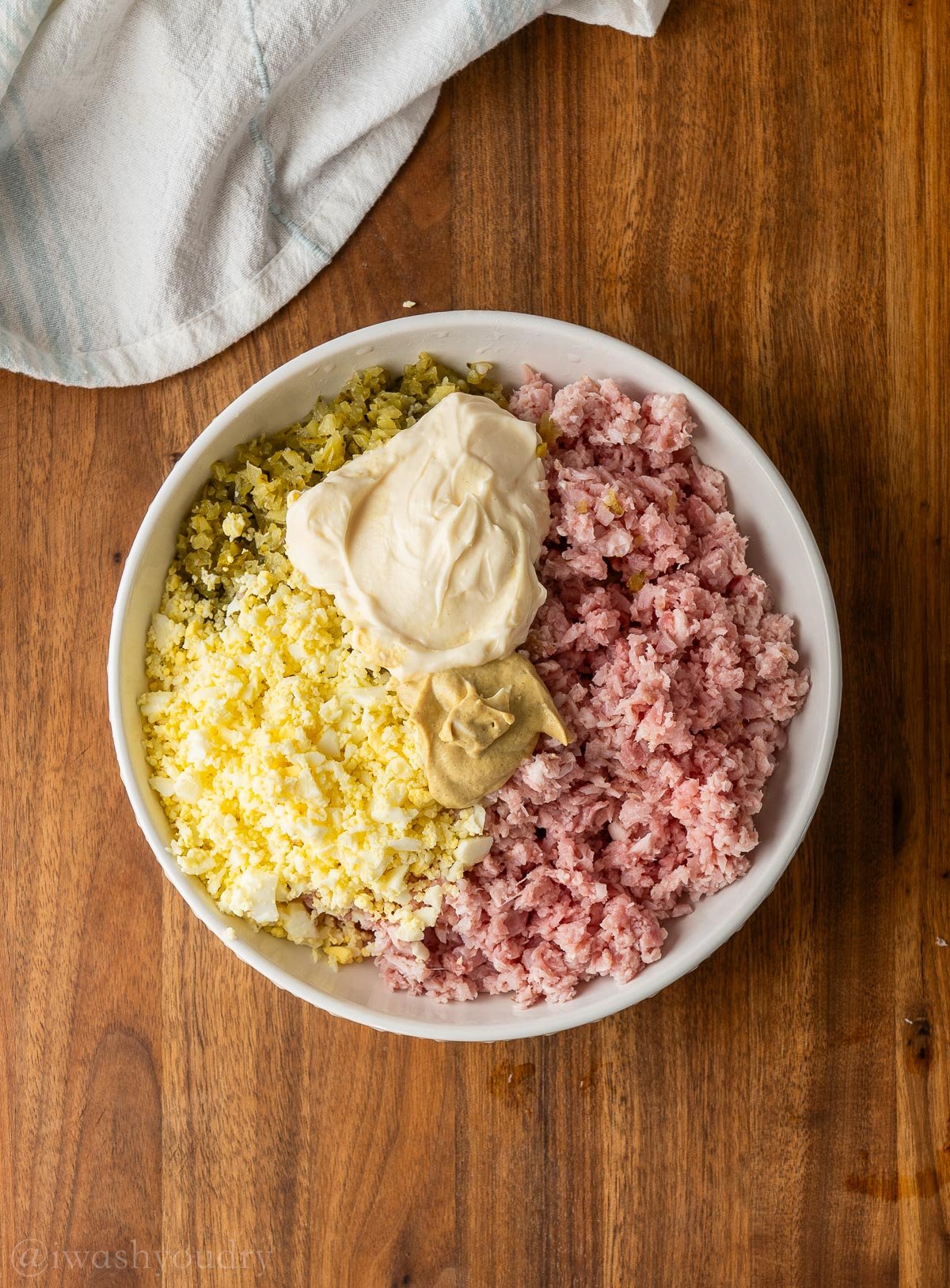 ingredients for chopped salad in a white bowl, topped with mayo and mustard.